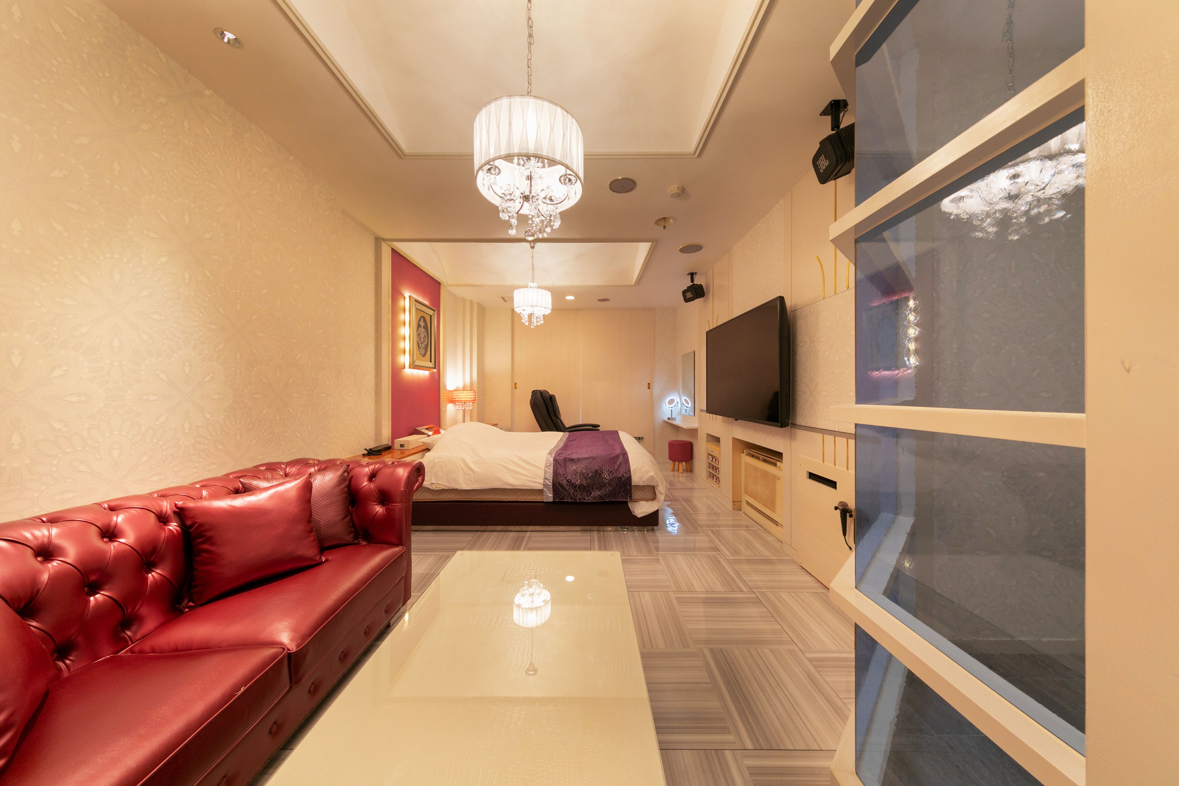 A modern hotel room with a red leather sofa, a large flat-screen TV, a glass enclosure, and a chandelier.