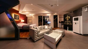 A modern hotel room with a sofa, a glass coffee table, and a bed with a patterned bedspread.