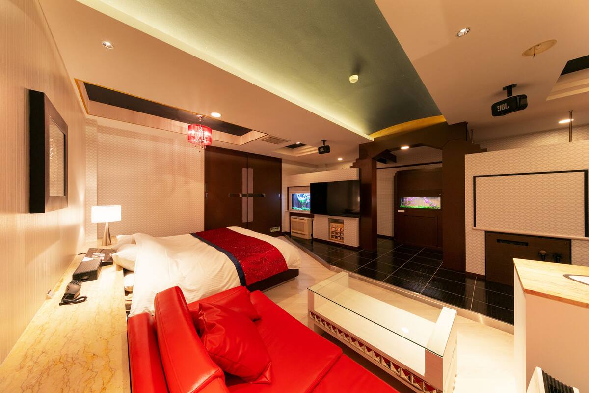 Une chambre d’hôtel moderne avec un grand lit, un canapé rouge, un bureau avec une lampe, une télévision et un mini-bar.