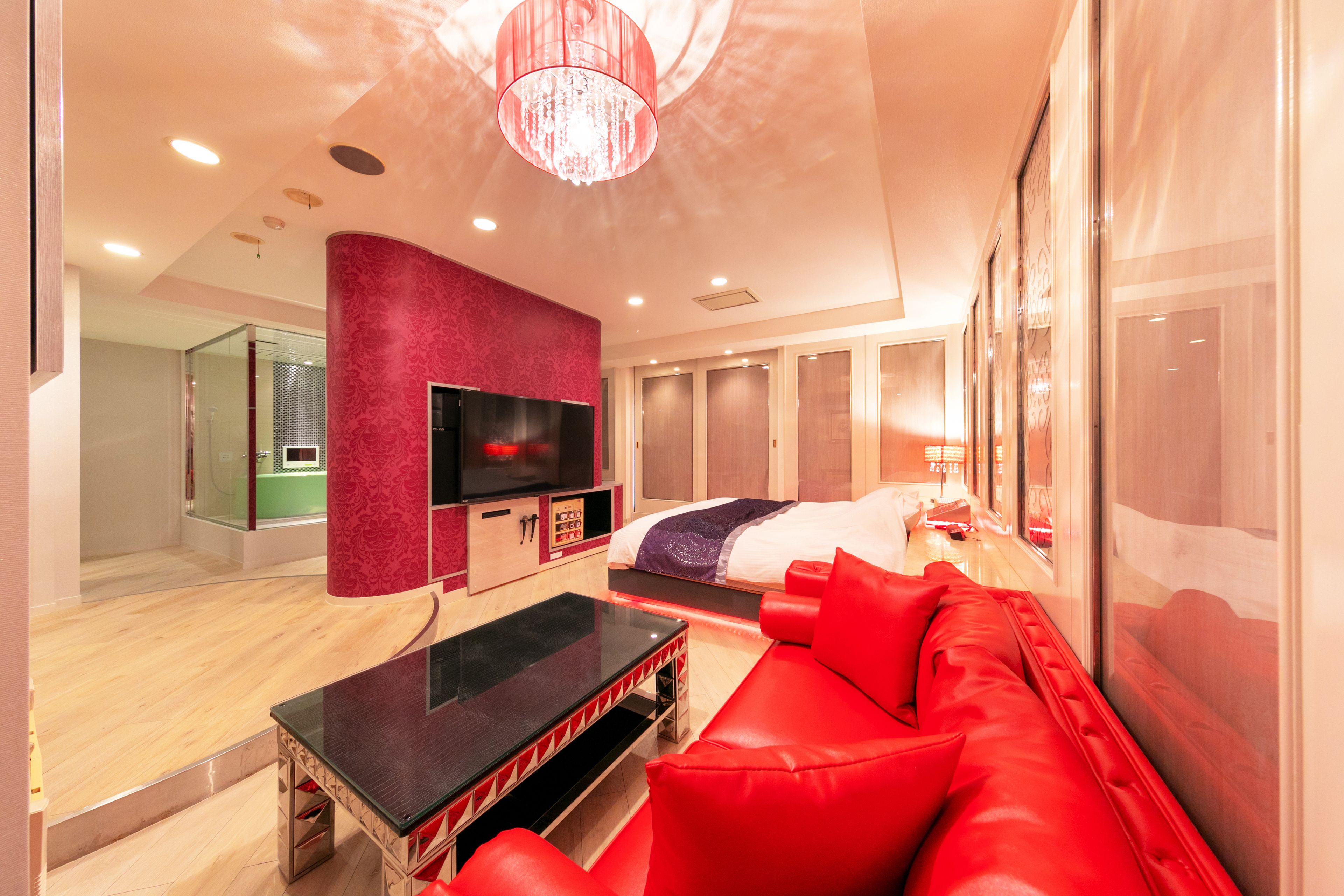 A modern hotel room with a red sofa, a flat-screen TV, a bed with a purple and white duvet, and a large mirror.
