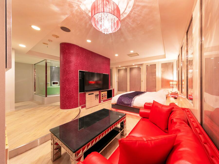 A modern hotel room with a red sofa, a flat-screen TV, a bed with a purple and white duvet, and a large mirror.