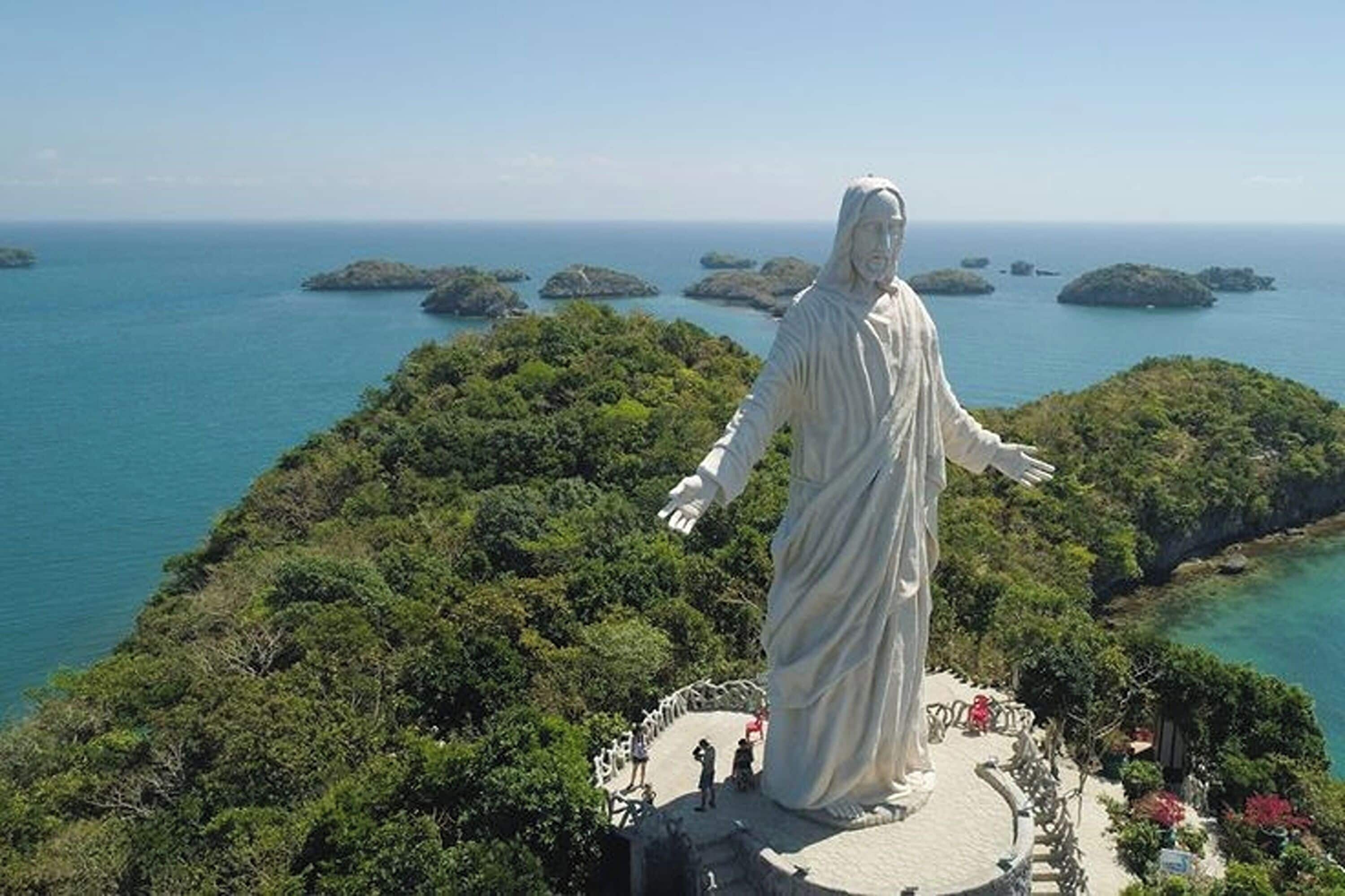 A statue of a robed figure with outstretched arms, surrounded by lush greenery and overlooking a coastal area with small islands in the distance.