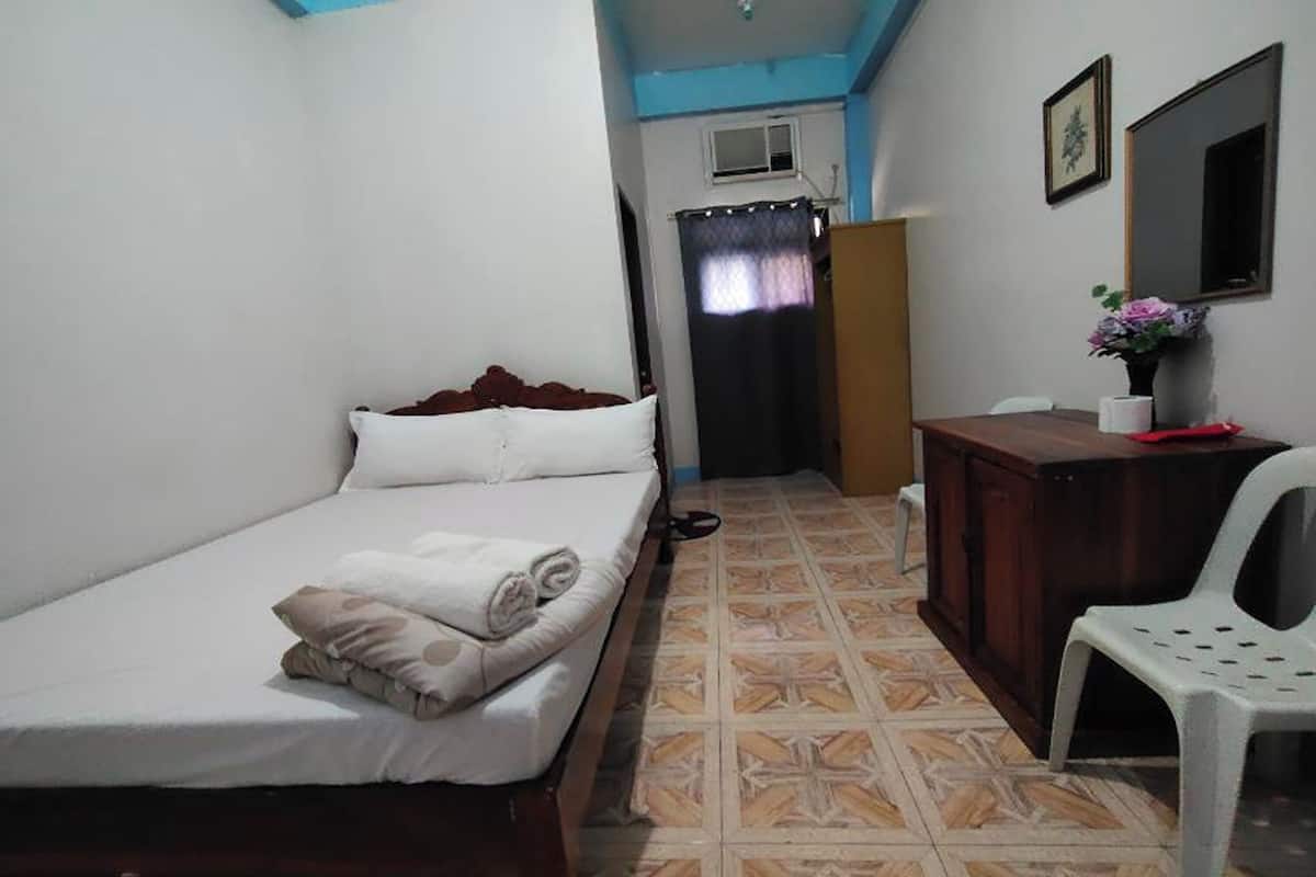 A small, simple room with a bed, a desk, a chair, and a window.