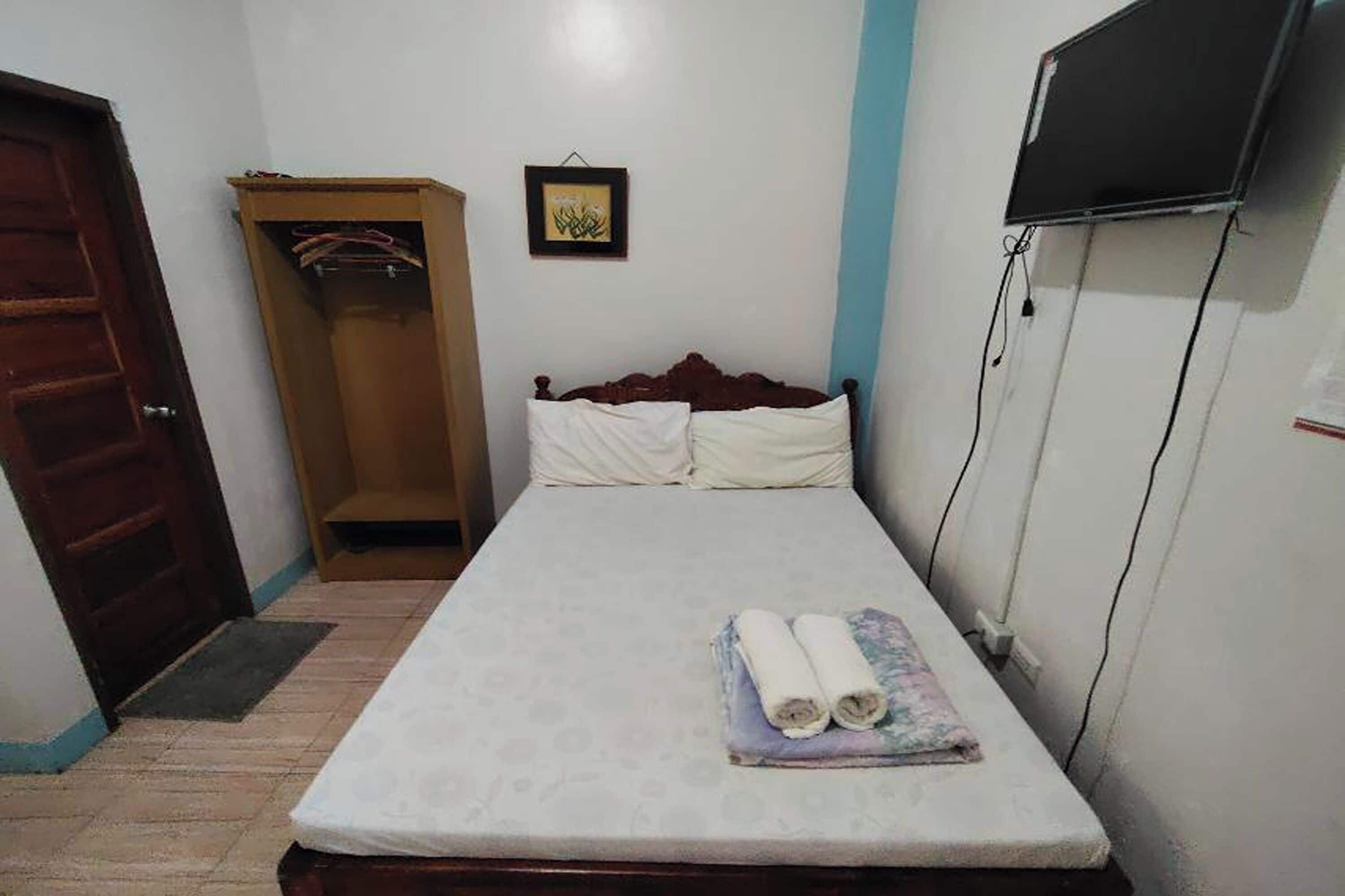 A small, neatly arranged bedroom with a single bed, a wooden wardrobe, a television mounted on the wall, and a framed picture above the bed.