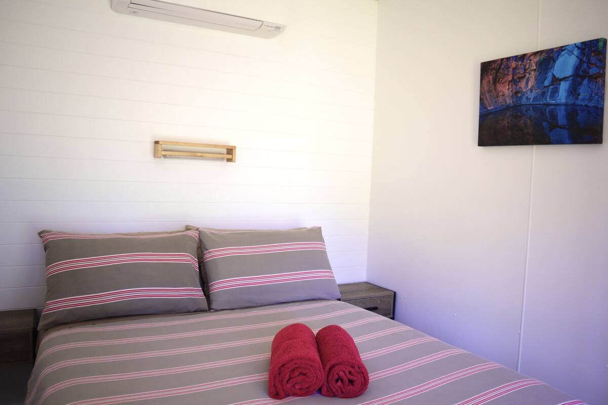 A neatly made bed with striped bedding and two pillows, a wooden nightstand, a wall-mounted light fixture, and a painting on the wall.