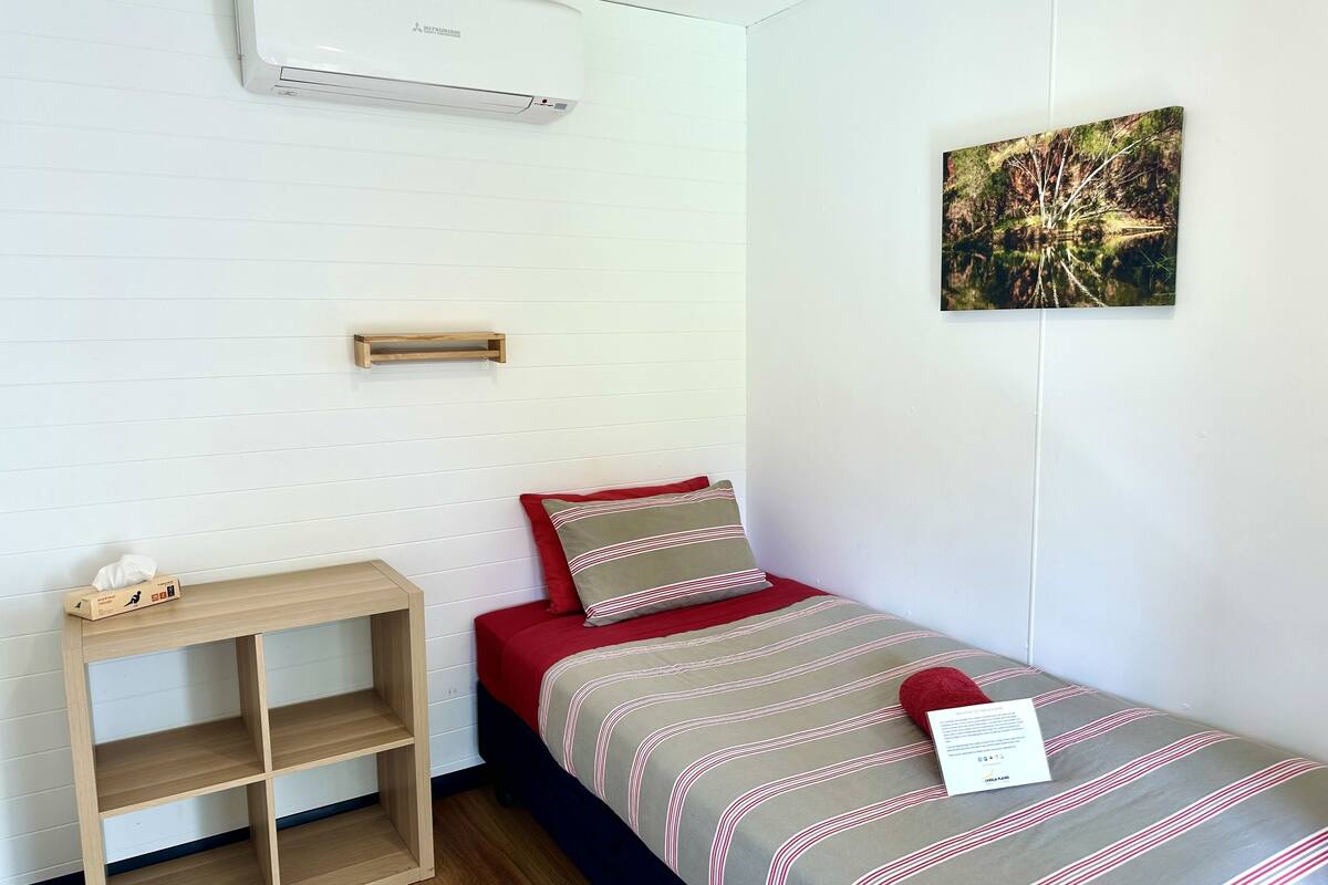 A small, neatly arranged bedroom with a bed, a wooden nightstand, a wall-mounted air conditioner, and a framed picture on the wall.