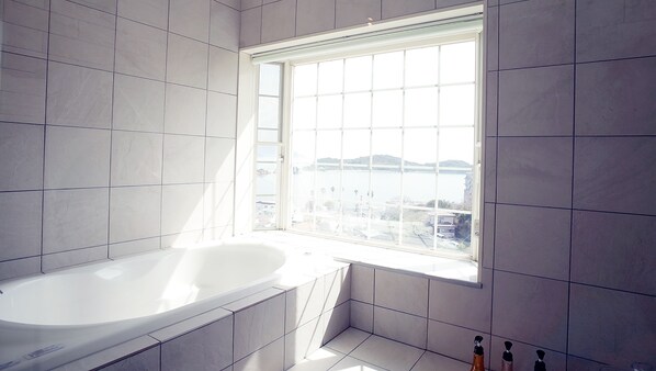 A modern bathroom with a large bathtub, a window with a view of a body of water and buildings, and tiled walls.