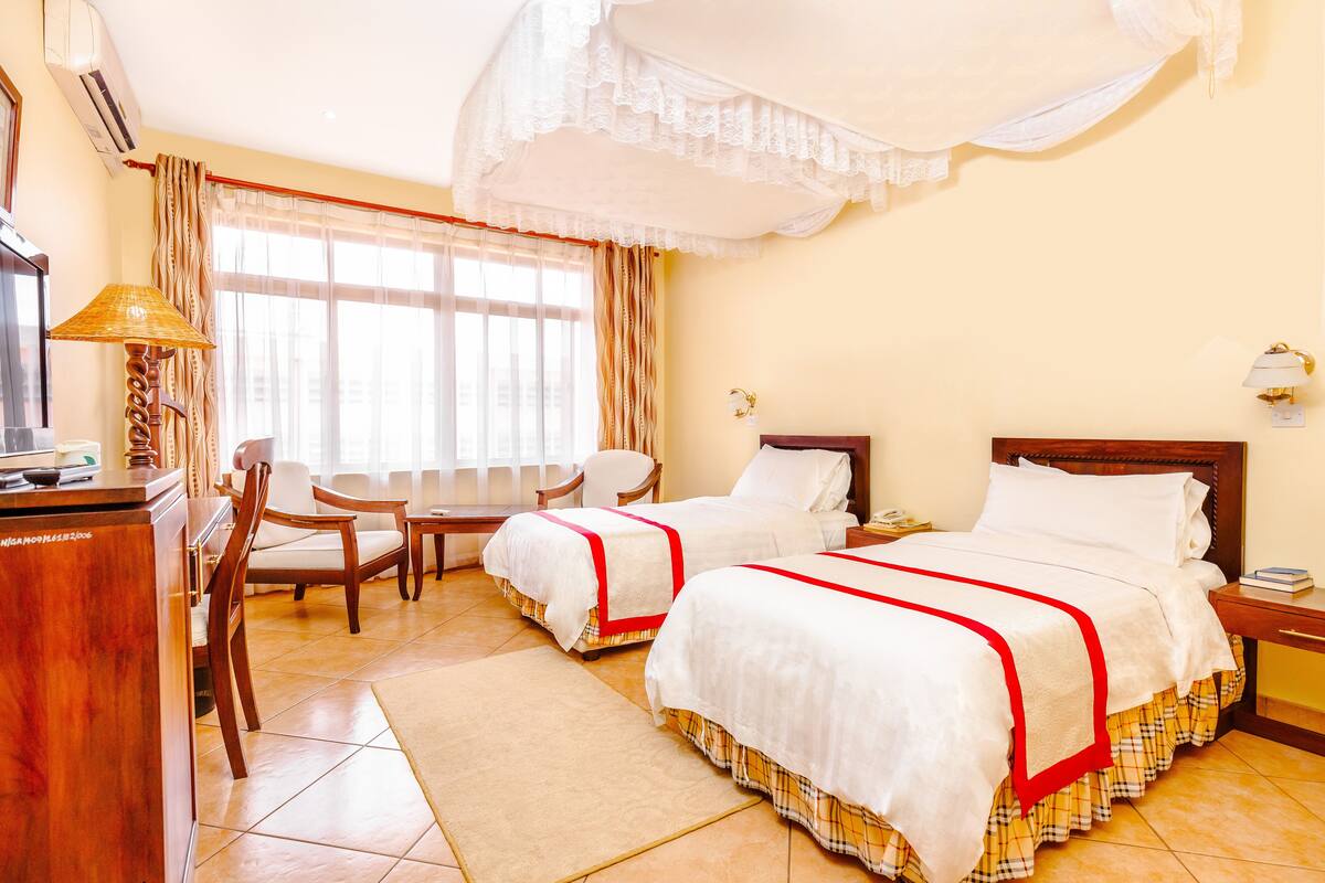A hotel room with two beds, a desk, a chair, a lamp, and a window with curtains.