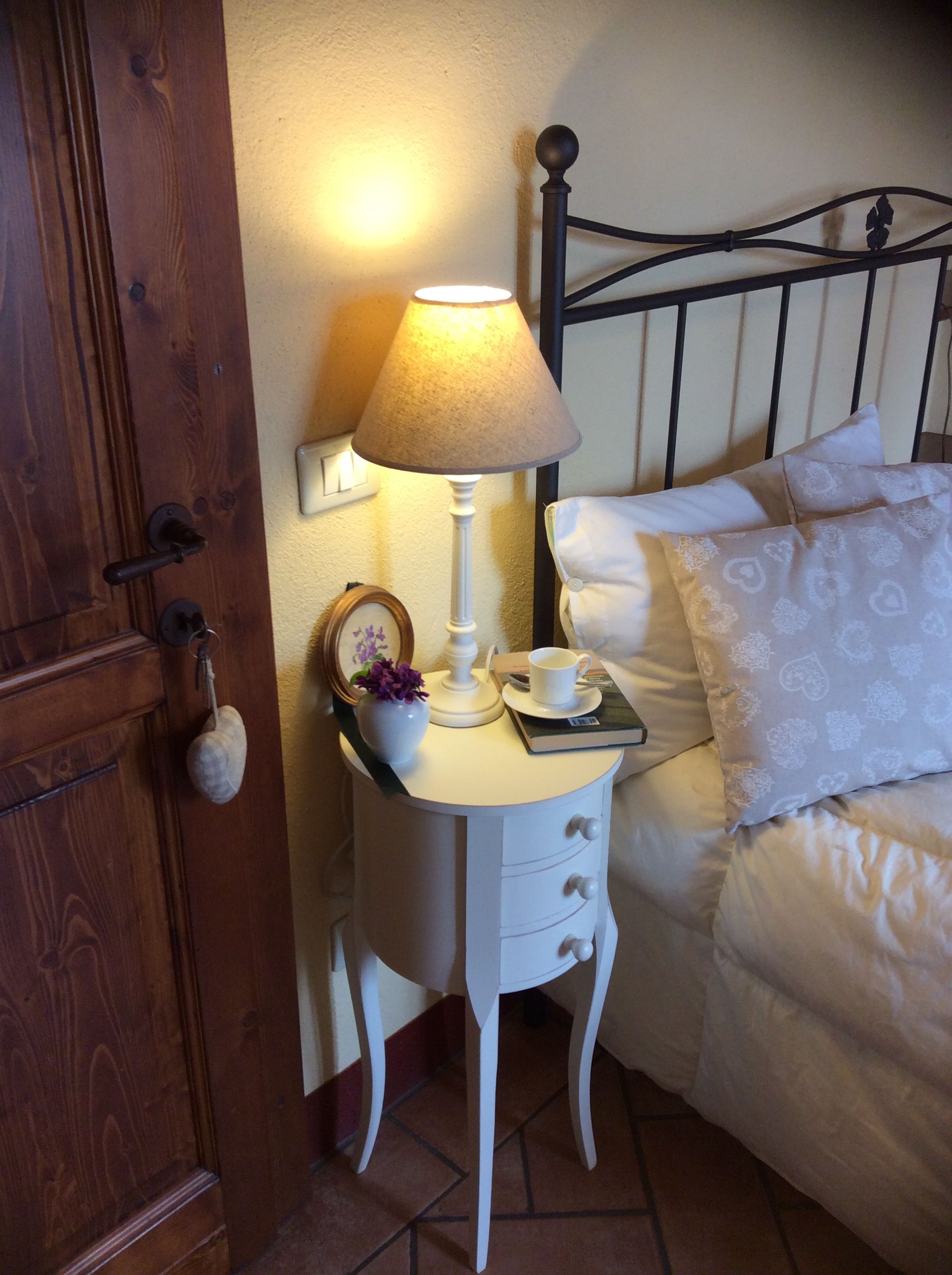 A neatly made bed with white linens and patterned pillows, a bedside table with a lamp, a cup, a saucer, and a small decorative item.