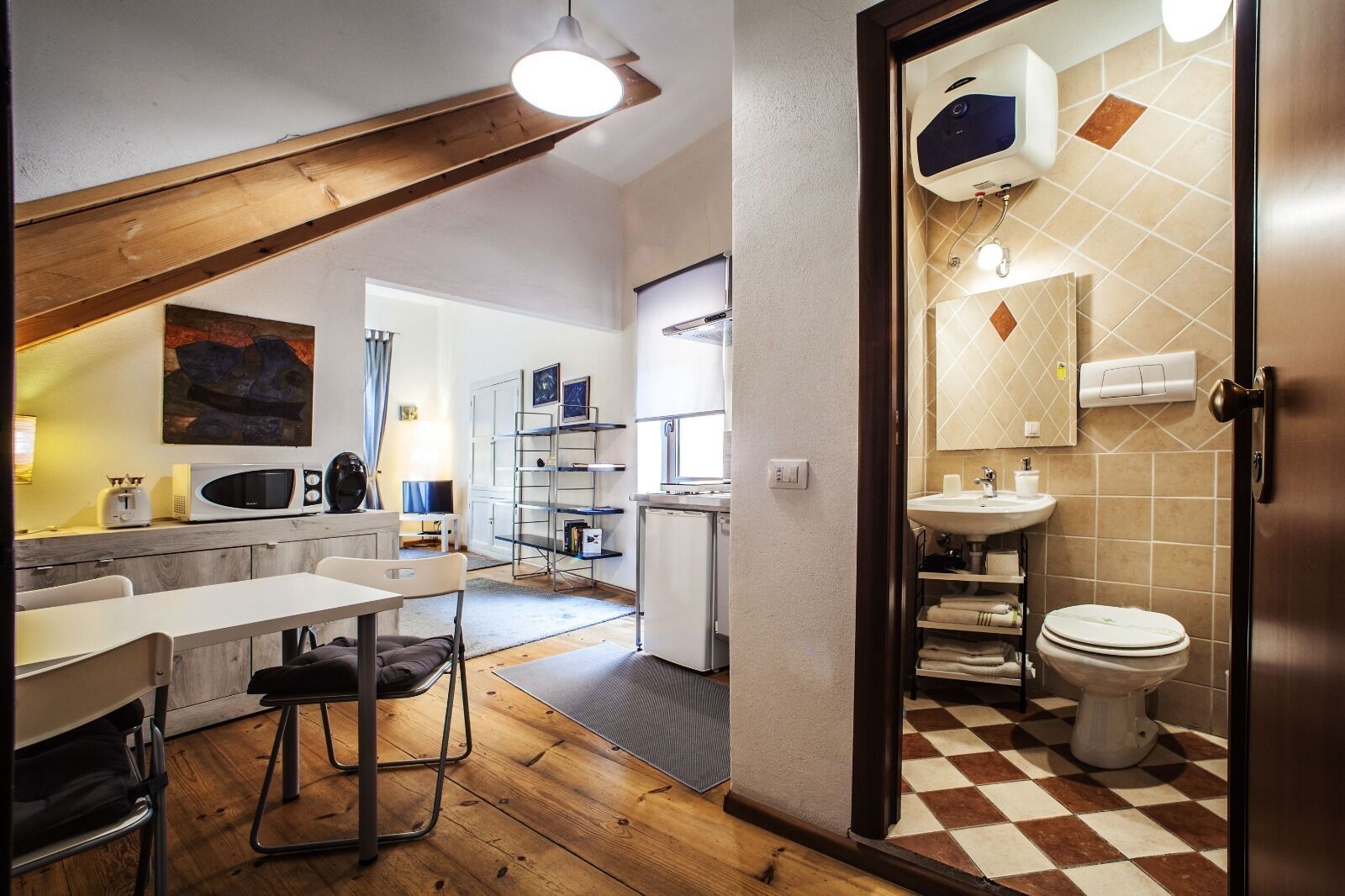 A compact kitchen with a microwave, toaster, and a small refrigerator. There is a dining area with a table and chairs. The bathroom has a toilet, sink, and a mirror with a towel rack.