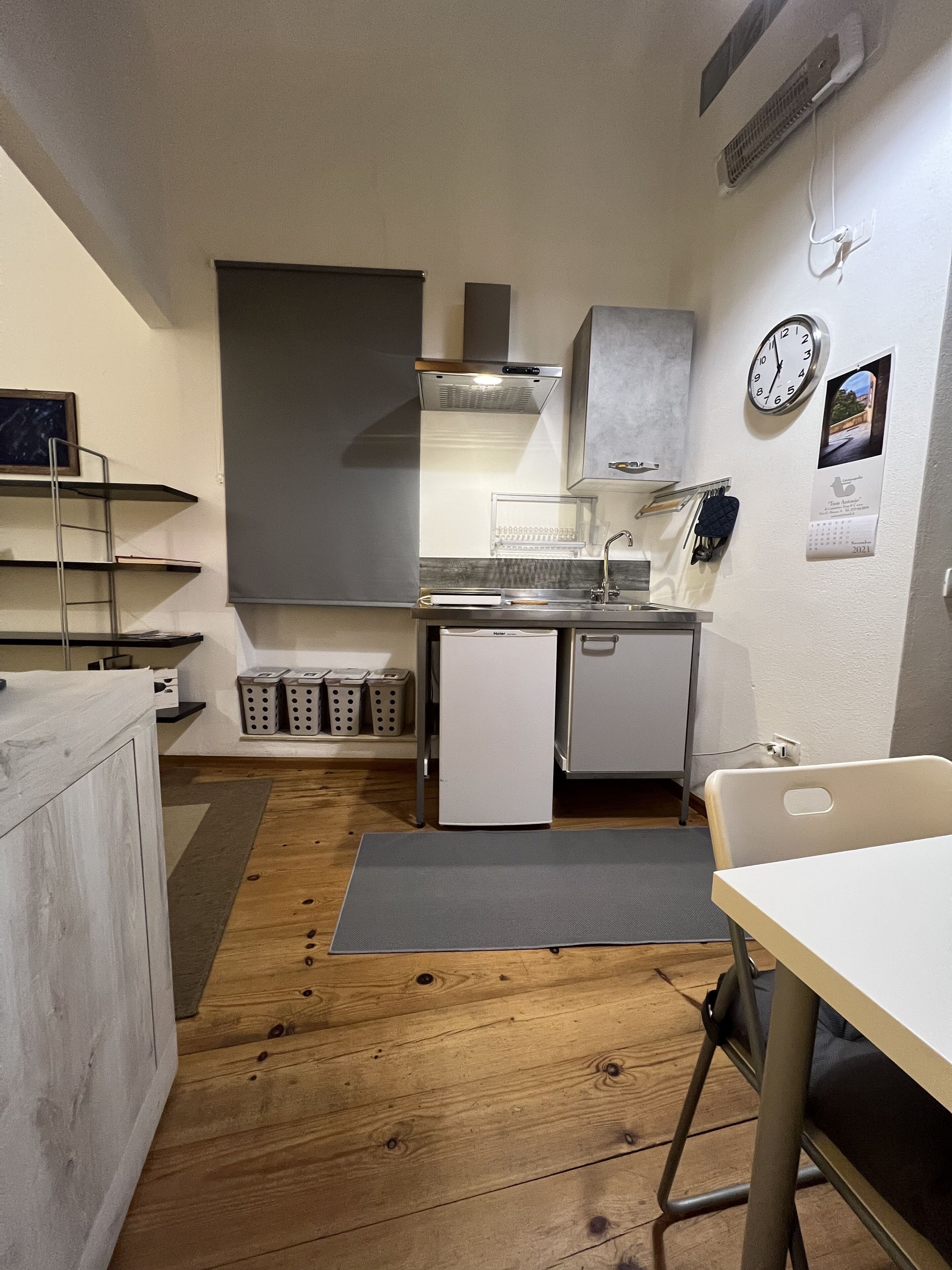 Une cuisine compacte équipée d’une cuisinière intégrée, d’un petit réfrigérateur et d’un évier. On y trouve des étagères et un égouttoir, ainsi qu’une horloge et un calendrier accrochés au mur.