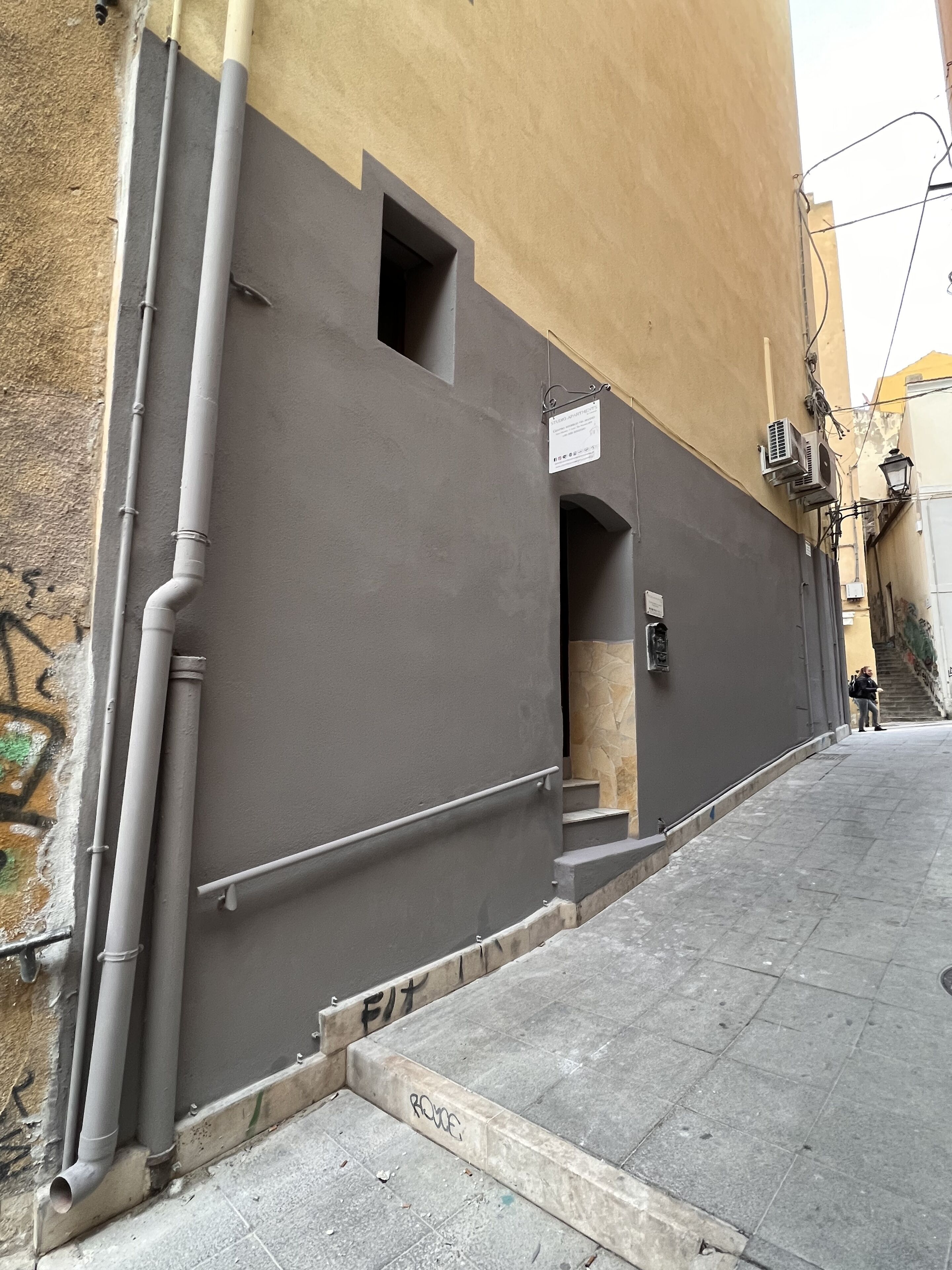 Une rue étroite bordée d’un immeuble à la façade grise, doté d’une petite fenêtre et d’un appareil de climatisation apparent.