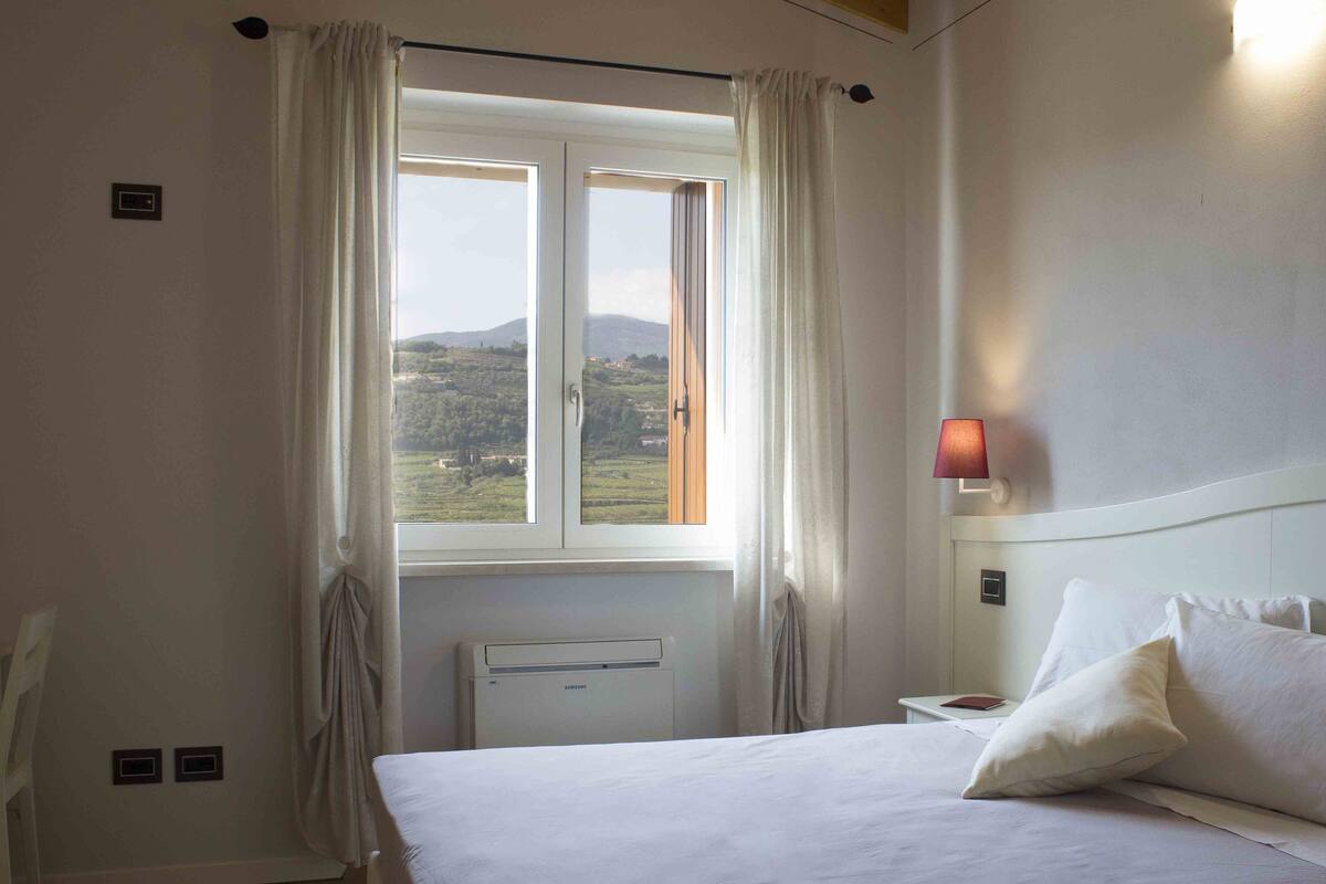 Une chambre d’hôtel avec un lit, une table de chevet, une lampe, une fenêtre avec des rideaux et une vue sur la campagne.