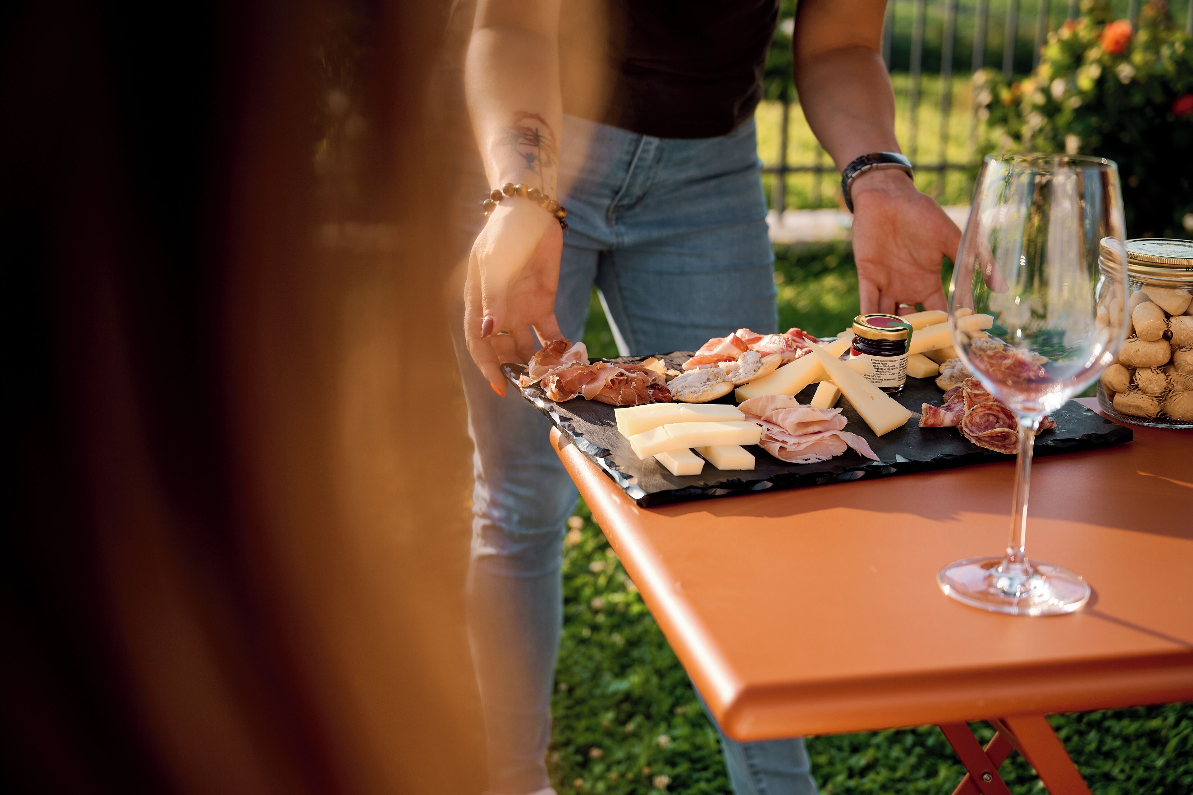 Eine Person serviert eine Platte mit Wurst- und Käsesorten an einem Tisch im Freien.