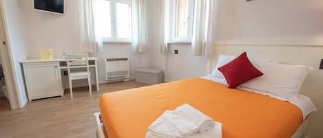 Une chambre d’hôtel avec un lit, un bureau et une chaise. Il y a une fenêtre avec des rideaux, un téléviseur à écran plat fixé au mur et un tableau au-dessus du lit.