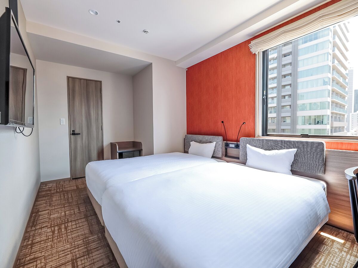 A hotel room with a large bed, a desk, a chair, a TV, and a window with a city view.