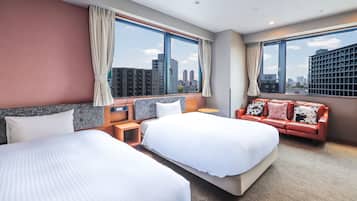Habitación de hotel con dos camas, un sofá, una mesita y grandes ventanales con vistas a la ciudad.