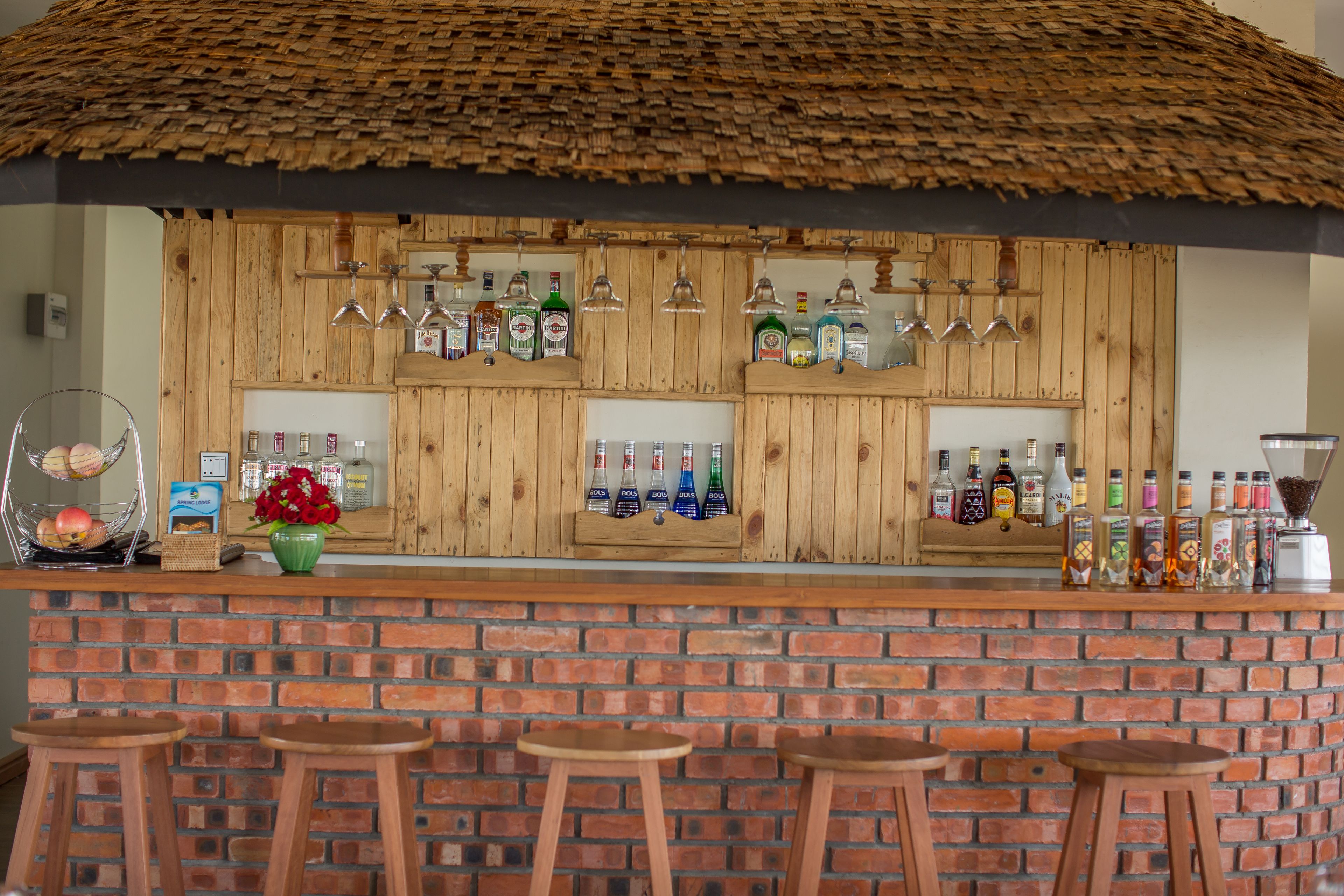 Una zona de bar con estantes de madera llenos de botellas y vasos, una pared de ladrillo y taburetes de madera.