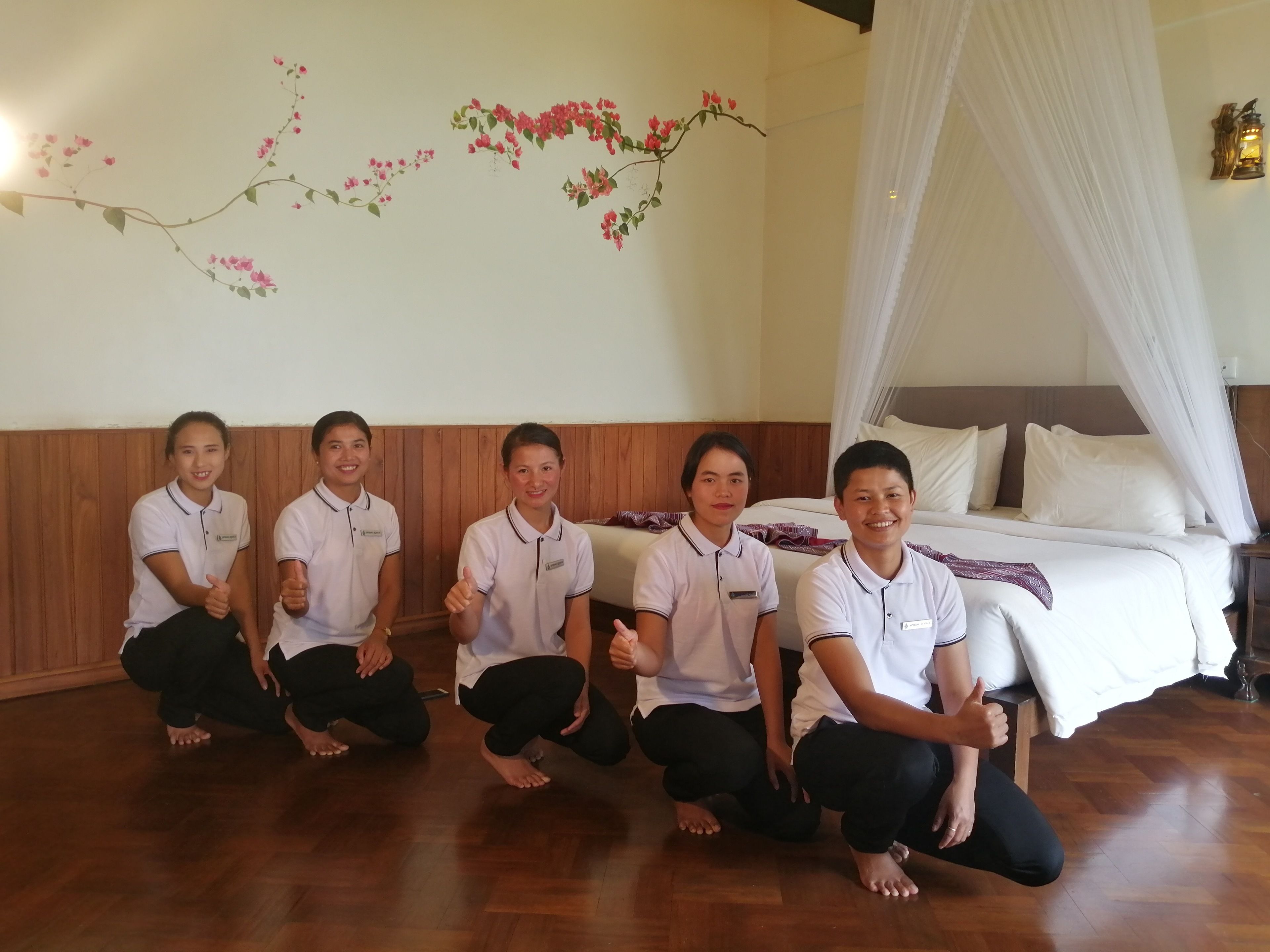 A group of people in a room with a bed, a canopy, and a wall with floral decorations.