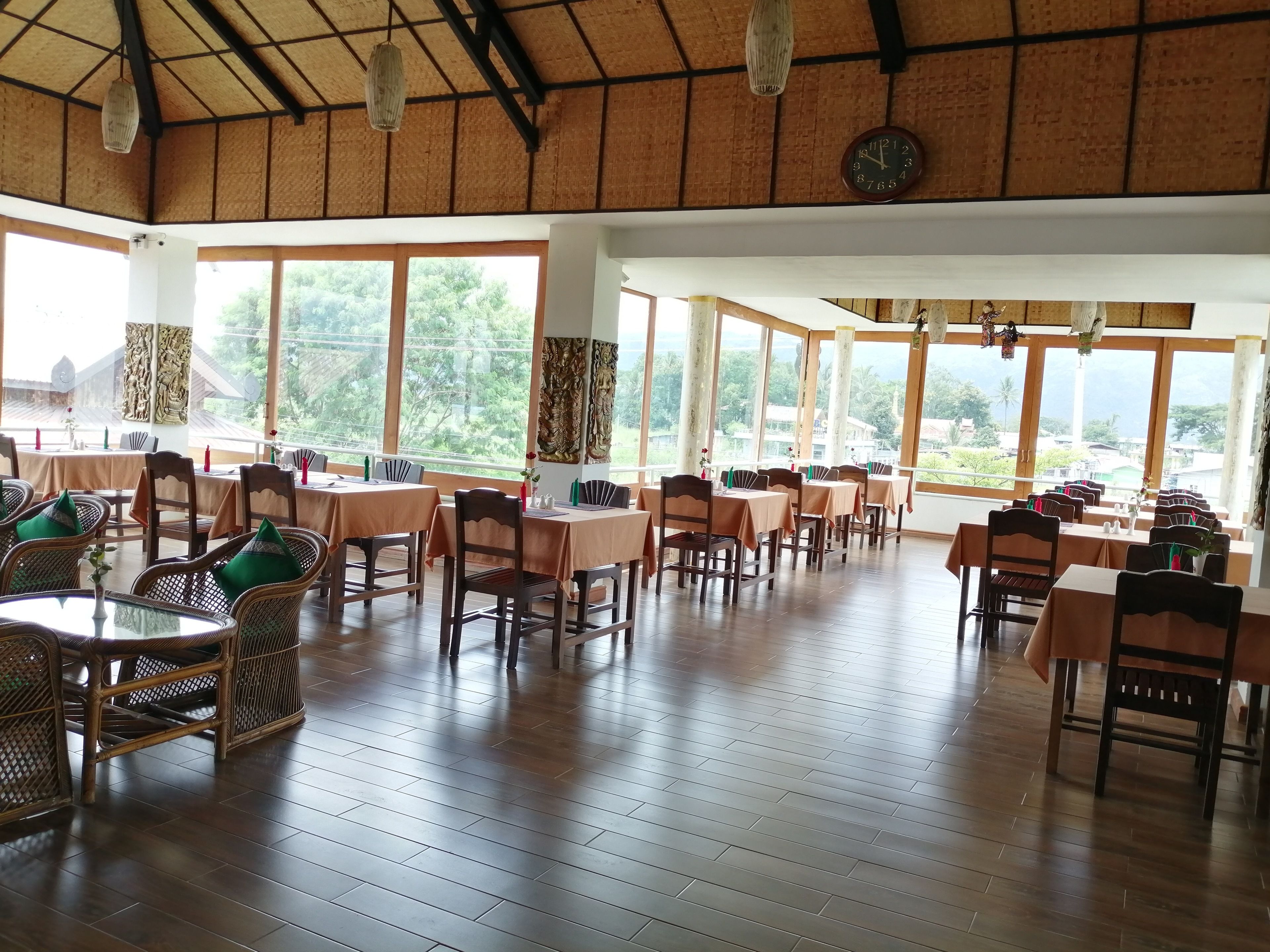 A spacious dining area with wooden tables and chairs, large windows offering a view of the outdoors, and a rustic ceiling design.