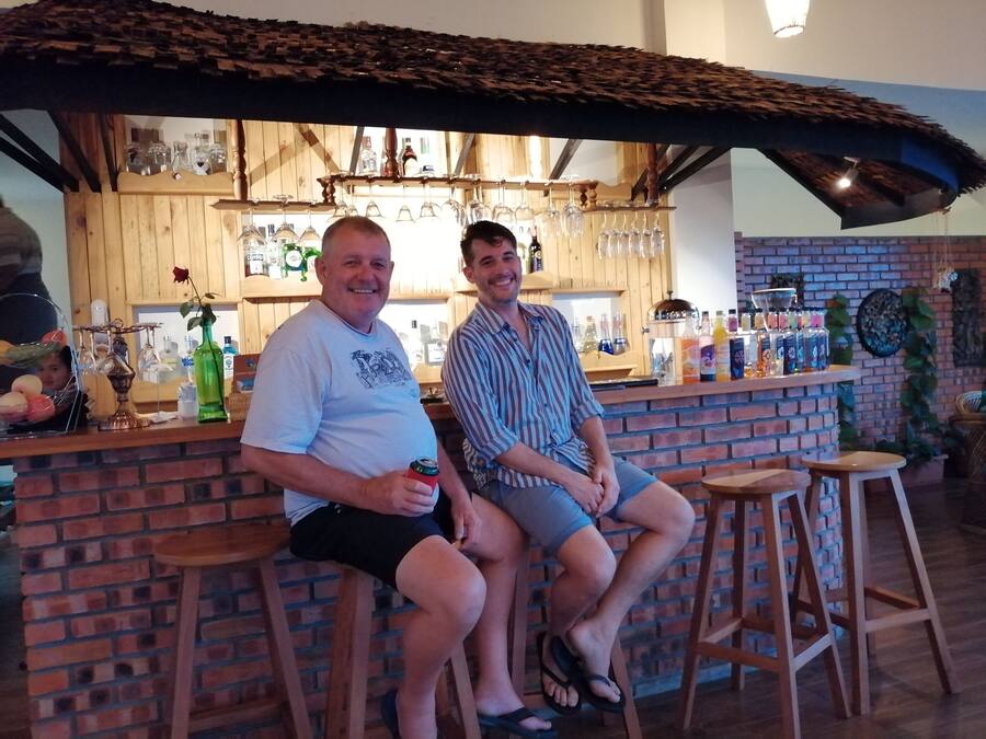 Dos personas sentadas en un bar con diseño rústico de madera, que presenta una pared de ladrillo y estantes con botellas y copas.