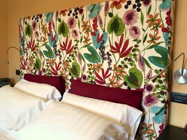 Habitación de hotel con cabecera tapizada con motivos florales, dos camas con ropa blanca y una mesita de noche con lámpara.