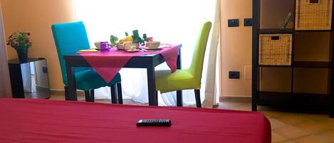 A hotel room with a bed, a table with chairs, a television, and a small cabinet with baskets.