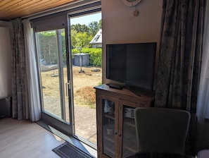 A room with a sliding glass door leading to a backyard, a television on a wooden cabinet, and a chair. - B&B de Zeemeeuw (Renesse)