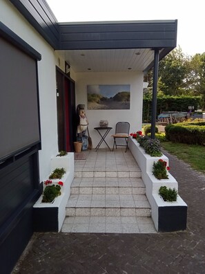 Steps lead to a covered porch with a table and chair, surrounded by lush greenery and a paved pathway.