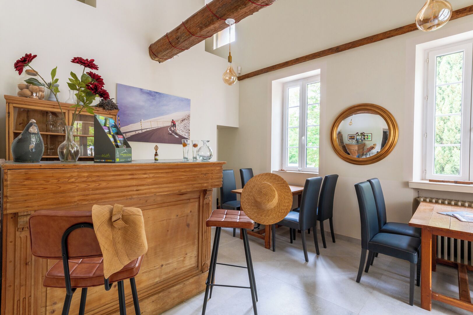 A dining area with a wooden bar, chairs, and a table. There is a painting on the wall and a window with a view of greenery.