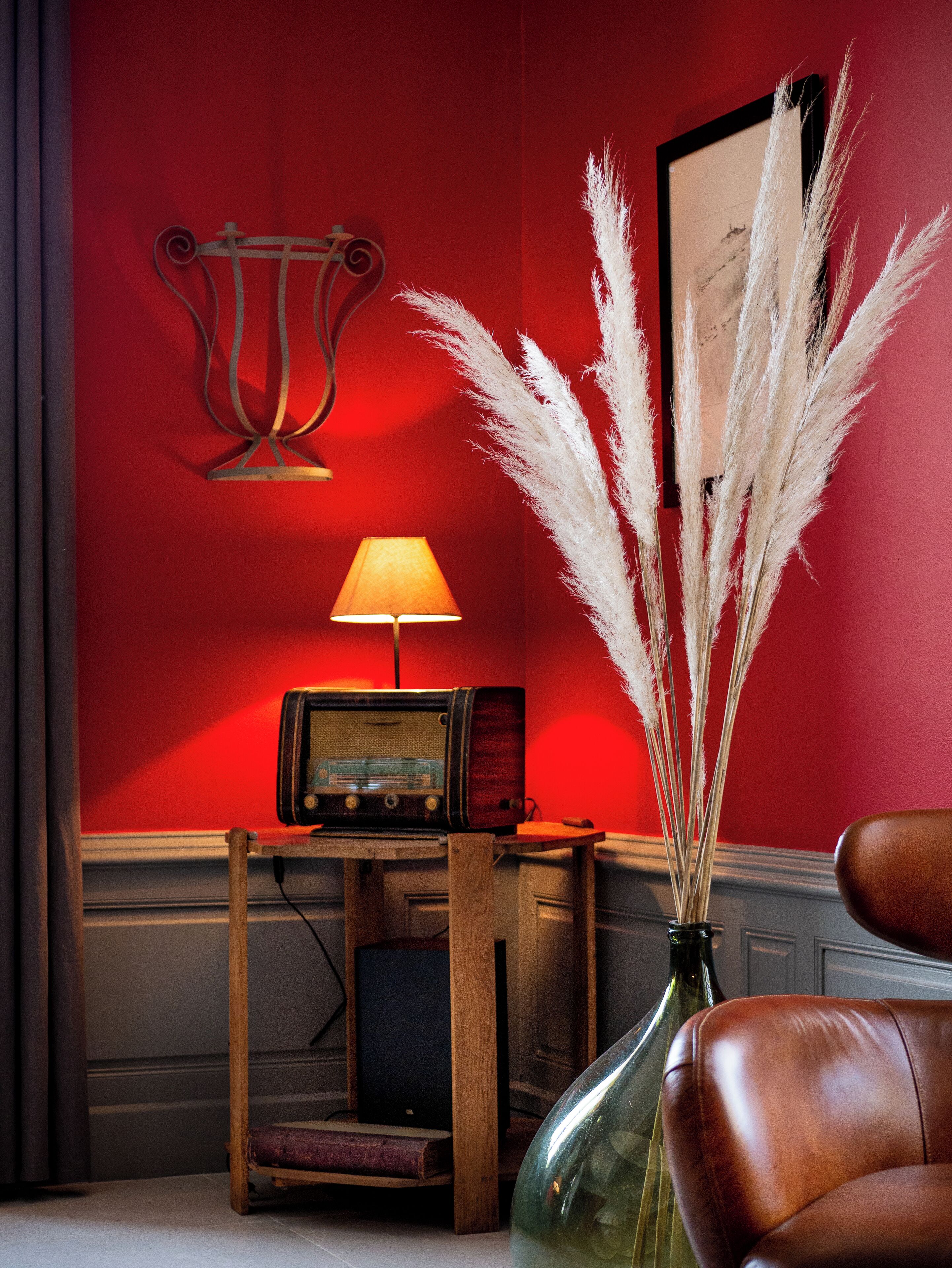 A room with a red wall, a vintage radio, a lamp, and a vase with pampas grass.