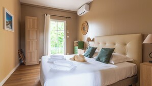 A neatly made bed with white linens, a hat, and towels, a bedside lamp, and a view of greenery through a window. - Domaine des Tilleuls (Malaucene)