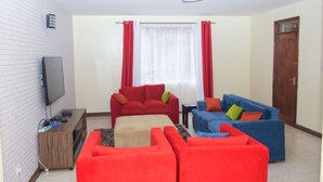 A living room with a flat-screen TV, a wooden entertainment unit, red and blue sofas, and a window with red curtains. - Cozy and Furnished 1 Bedroom Apartment (Nairobi)