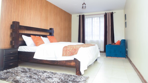 A bedroom with a wooden headboard bed, a blue sofa, and a large window with curtains. - Cozy and Furnished 1 Bedroom Apartment (Nairobi)