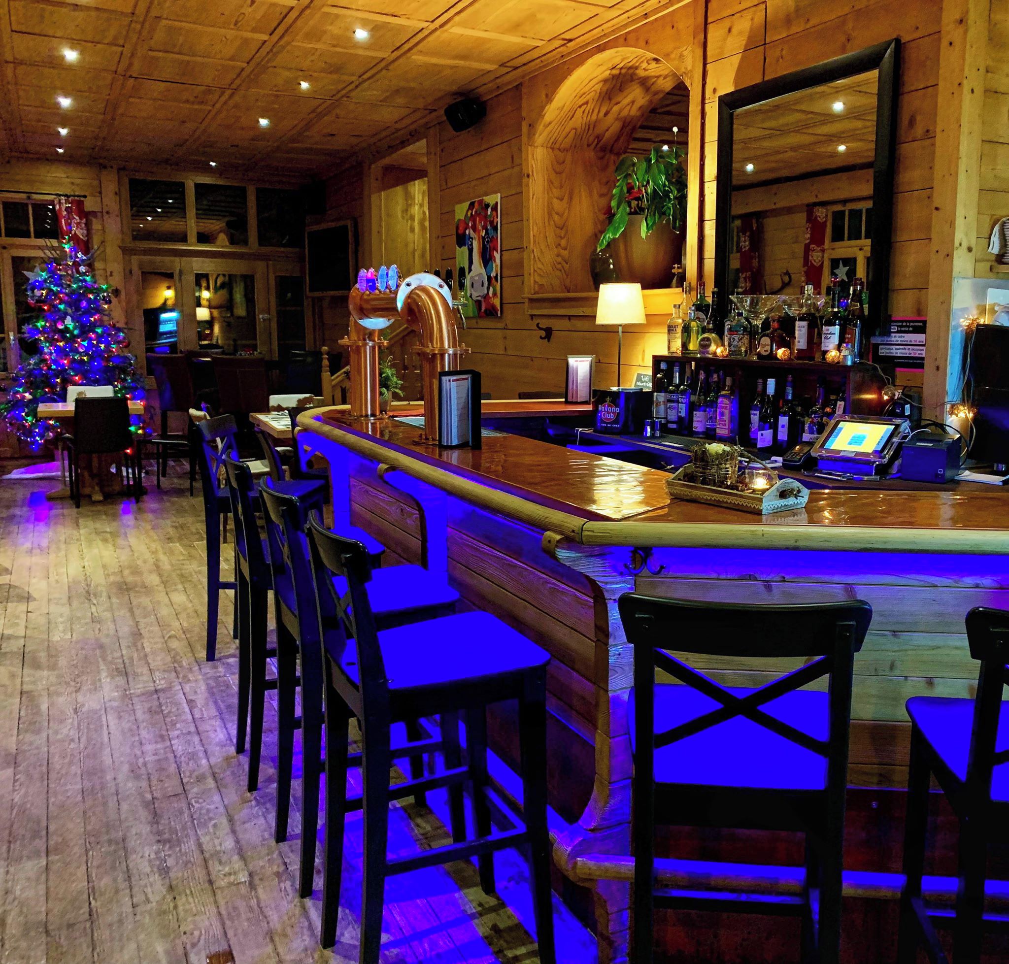 A bar area with wooden bar stools, a Christmas tree, and a variety of bottles on shelves.