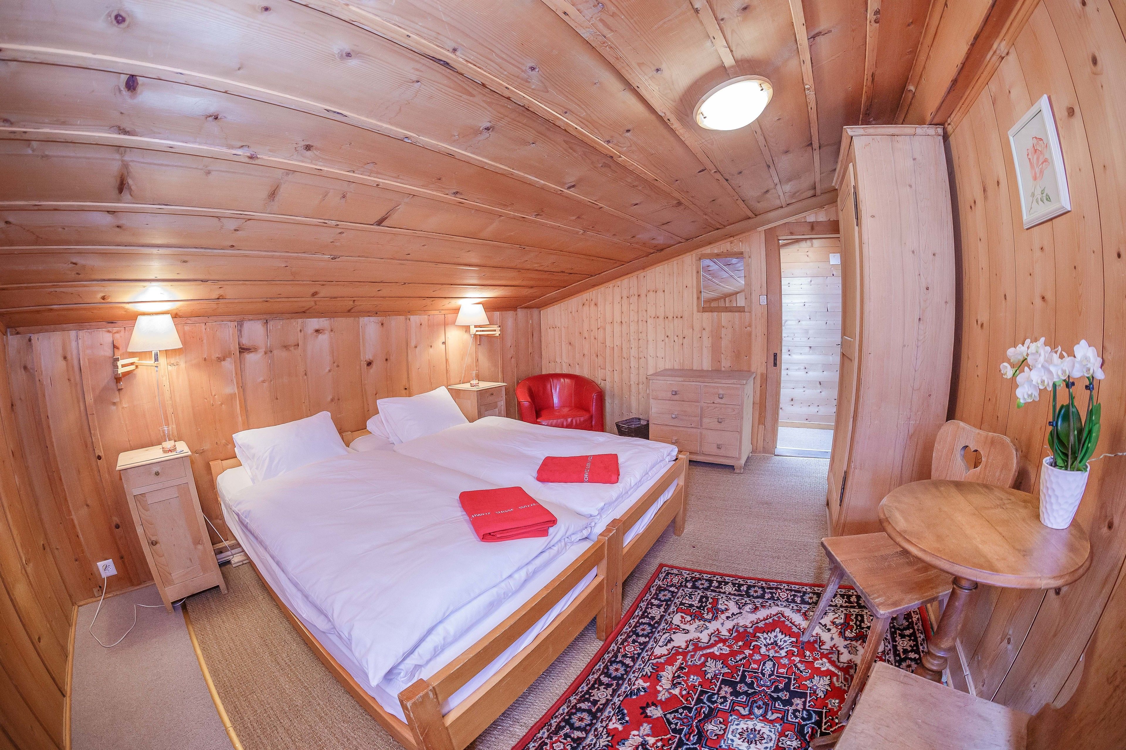 A wooden cabin room with a bed, a red chair, a small table, and a nightstand.