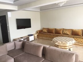 Una sala de estar moderna con un sofá color beige, un reposapiés estampado y un televisor de pantalla plana grande montado en la pared.