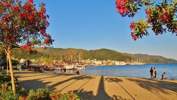 A coastal town with a sandy beach, boats docked at the harbor, and a backdrop of hills. - George Dragon Beach Hotel (Marmaris)