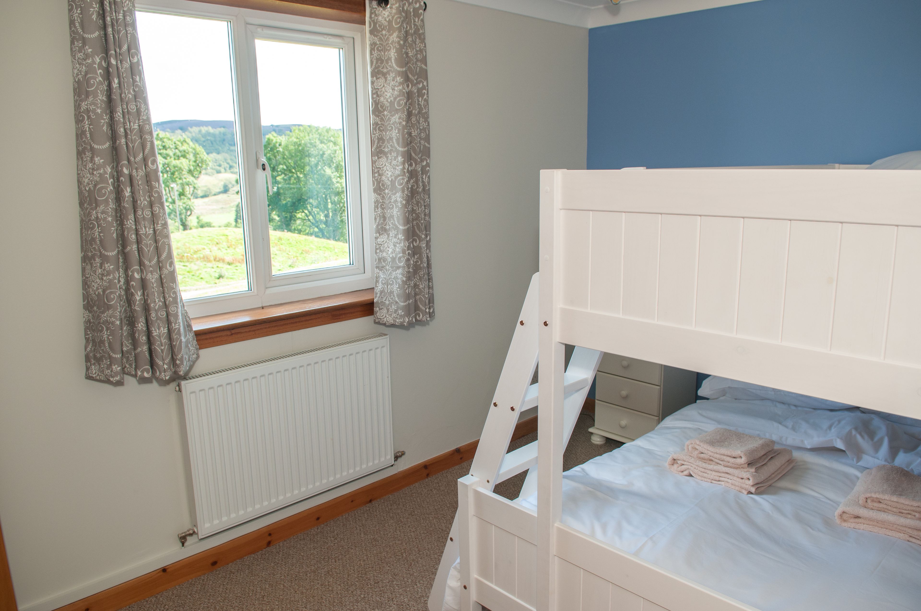 A bedroom with a bunk bed, a window with curtains, a radiator, and a view of greenery outside.