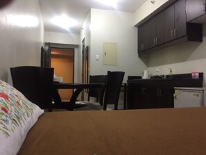 A compact room with a bed, a desk, a chair, a small kitchenette, and a door leading to another room.