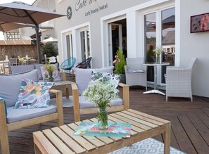 Un área de estar al aire libre con mesas y sillas de madera, un florero con flores y un cartel que dice 'Café Resto Hotel'.