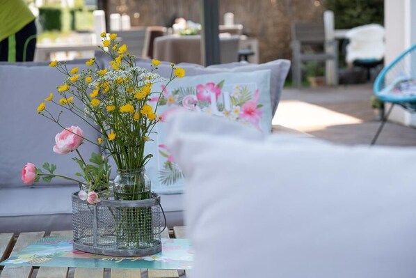 Una mesa con un arreglo floral en un florero de vidrio, un cojín estampado con flores y una silla al fondo.