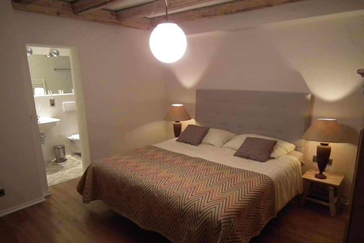 A bedroom with a large bed, two bedside lamps, a headboard, and a visible bathroom in the background.