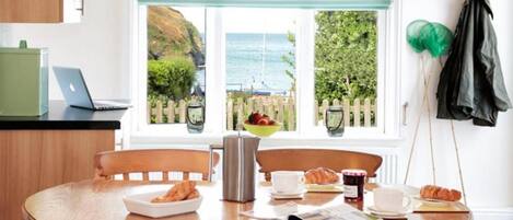 A dining table with breakfast items, a laptop, and a view of the sea through a window.