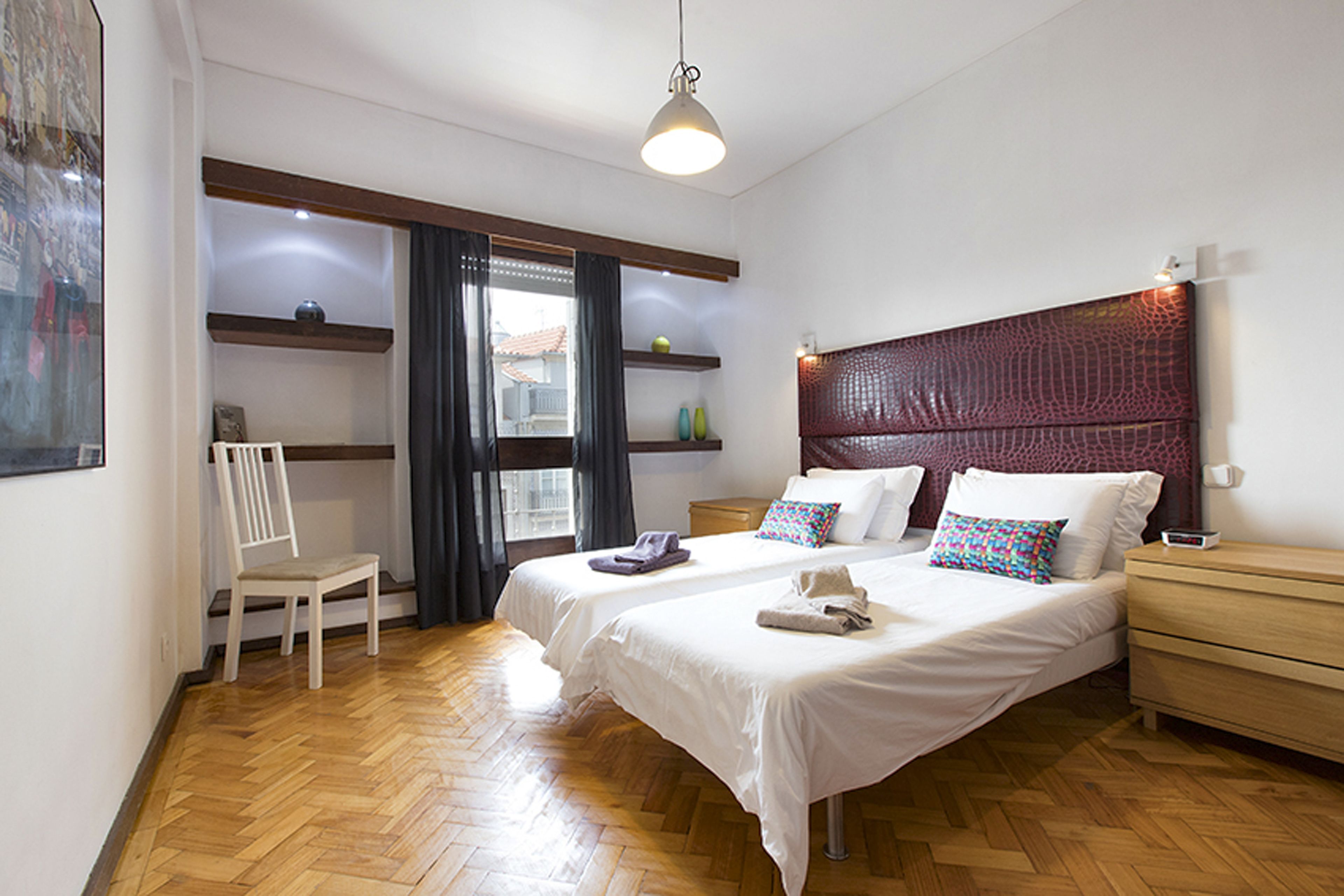 Un dormitorio con dos camas, suelo de madera, una silla, una mesita de noche y una ventana con cortinas.