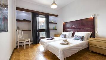Un dormitorio con dos camas, suelo de madera, una silla, una mesita de noche y una ventana con cortinas.