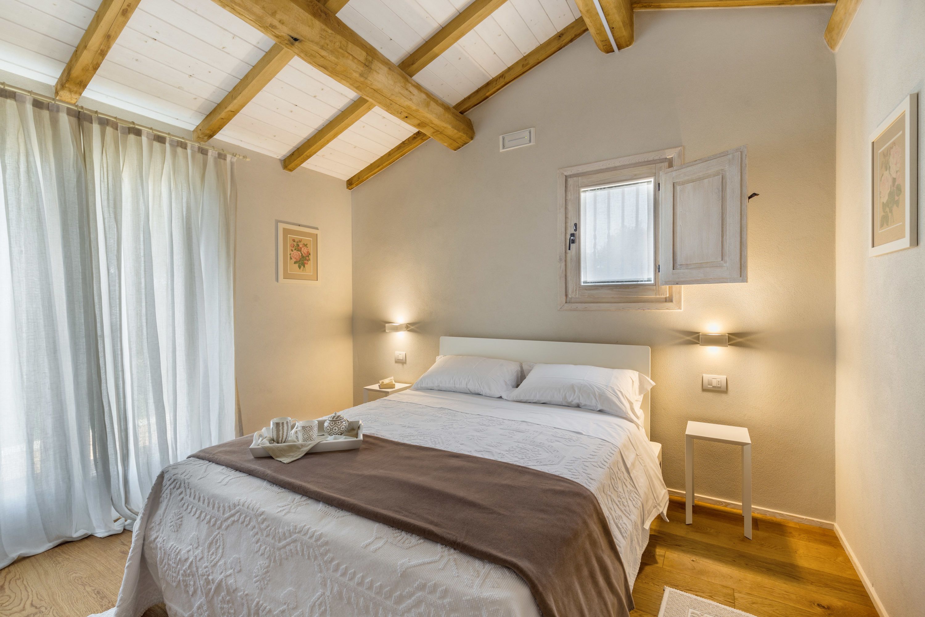 Un dormitorio con una cama grande, vigas de madera en el techo, una ventana con marco blanco y una mesita de noche con una lámpara.