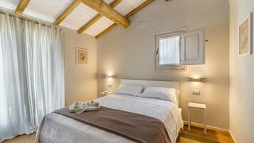 Un dormitorio con una cama grande, vigas de madera en el techo, una ventana con marco blanco y una mesita de noche con una lámpara.