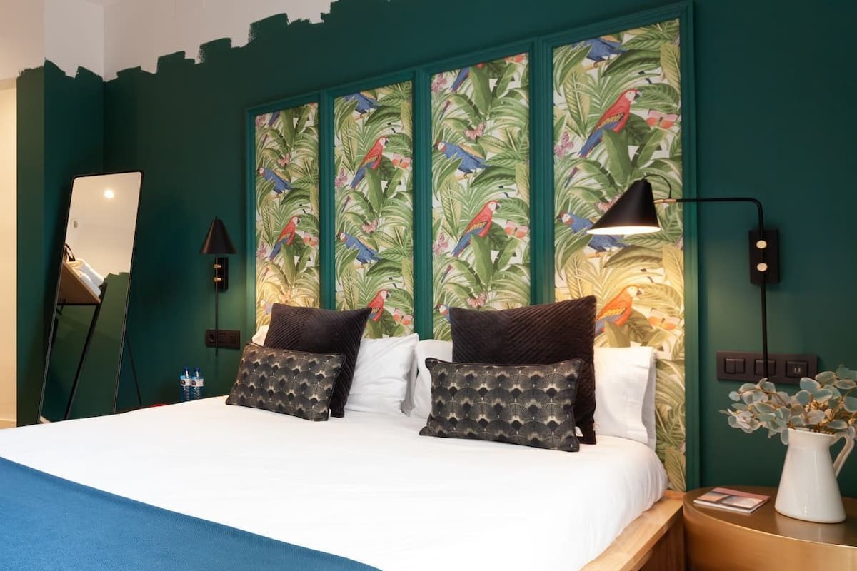 Ein Hotelzimmer mit einem großen Bett, zwei Kissen, einem Nachttisch, einer Lampe, einem Spiegel und einem Wandgemälde im tropischen Stil.