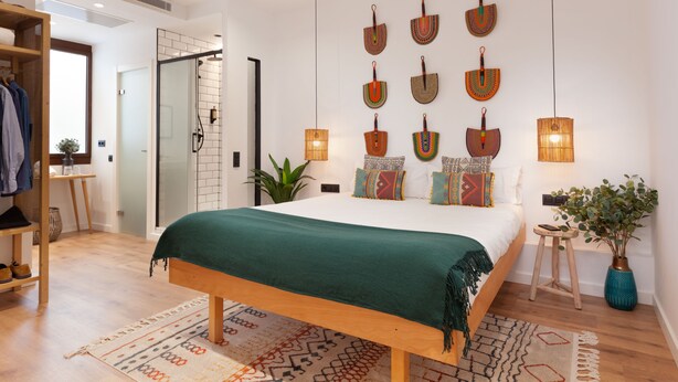A modern bedroom with a large bed, a wooden bench, a small stool, a potted plant, and a wall-mounted shelf. - Casa Vaganto (Barcelona)
